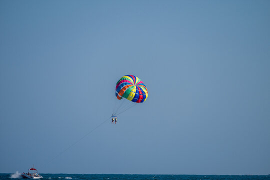 A motorboat tows a colorful rainbow parasail carrying two people soaring high above the open sea under a clear blue sky.