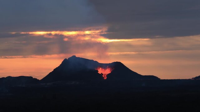 Volcanic eruption with glowing magma bubbling from crater in Fagradalsfjall silhouetted against a dramatic sunset sky in Iceland