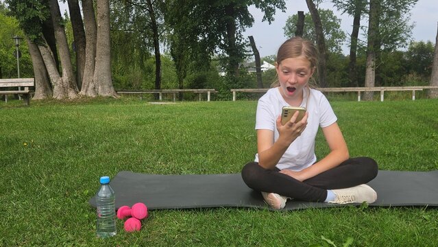 Young girl in a white t‑shirt and black leggings sits cross‑legged on a dark grey yoga mat in a park. She looks excited and surprised while holding her smartphone up, mouth open in an expressive react