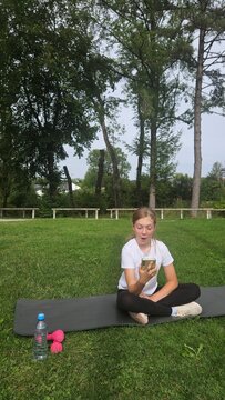 Young girl in a white t‑shirt and black leggings sits cross‑legged on a dark grey yoga mat in a park. She looks excited and surprised while holding her smartphone up, mouth open in an expressive react