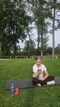 Young girl in a white t‑shirt and black leggings sits cross‑legged on a dark grey yoga mat in a park. She looks excited and surprised while holding her smartphone up, mouth open in an expressive react