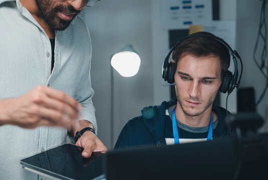 Technology IT Programmer Working At Computer With Headphones While Colleague Uses Tablet In Modern Office, Software Development Team Collaboration, Coding, Technical Support And Digital Business.