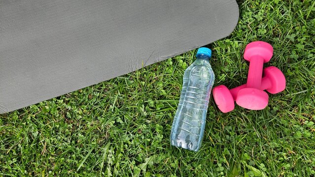 Close‑up outdoor shot showing a dark grey fitness mat partially in frame on the left, its smooth textured surface angled diagonally. On green grass in the foreground &mdash; a transparent plastic water bott