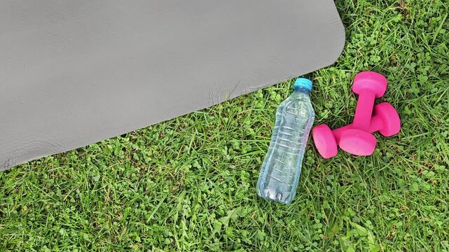 Close‑up outdoor shot showing a dark grey fitness mat partially in frame on the left, its smooth textured surface angled diagonally. On green grass in the foreground &mdash; a transparent plastic water bott