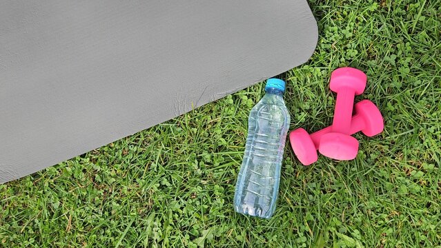 Close‑up outdoor shot showing a dark grey fitness mat partially in frame on the left, its smooth textured surface angled diagonally. On green grass in the foreground &mdash; a transparent plastic water bott