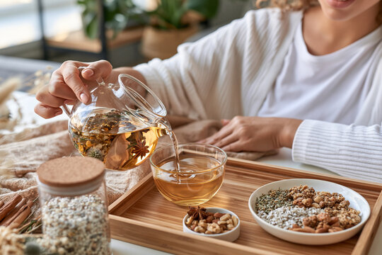 Woman pours warm herbal tea into a glass bowl. Her hands show calm care as she serves the drink. Wooden tray holds nuts, grains, and dried fruit. Soft lighting enhances cozy, health-focused atmosphere