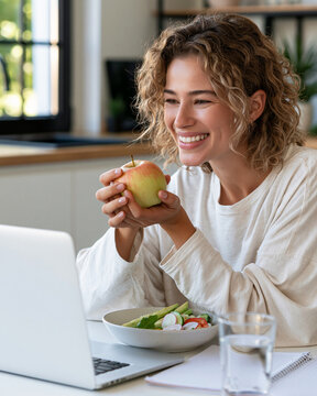 Woman smiles warmly, holding an apple. She eats healthy, seated at a laptop. Green salad sits beside her, with water. Soft daylight streams through window behind. Ideal for wellness, remote work