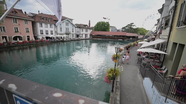 Riverside caf&eacute;s, flower-lined walkway and historic buildings beside the turquoise Aare River in Thun.