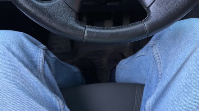 First-person view of driver legs in light denim jeans sitting behind car steering wheel. Feet positioned near vehicle pedals on dark floor mat.