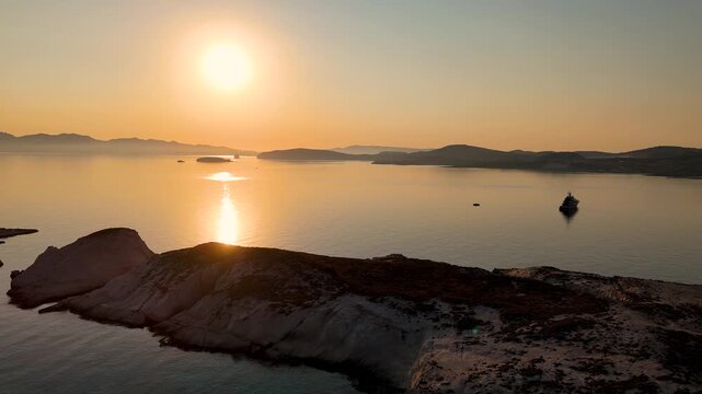 Aerial drone view flying over the aegean sea off the jagged coast of the island Milos in Greece during sunrise. The sun can be seen in the sky above the unique rocky landscape and ocean water. 