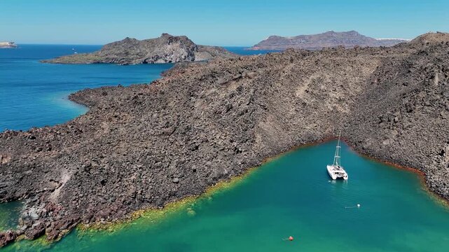 Drone ascends over the volcanic hot springs near Santorini, revealing rugged lava formations surrounded by turquoise Aegean Sea water and distant islands under bright Mediterranean sunlight.