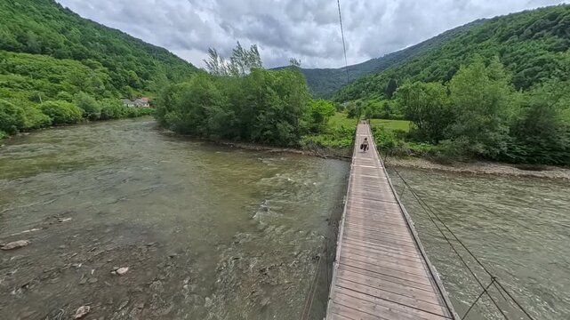 First-person POV perspective walking across a narrow wooden suspension footbridge spanning over a wide, fast-flowing mountain river toward the green forest shore.