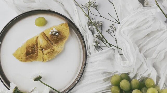 croissant,golden brown pastry,baked goods,flaky pastry,layered crust,browned surface,round plate,dark rim,white flower,yellow center,tiny blossoms,floral stem,branching stem,white fabric,draped fabric