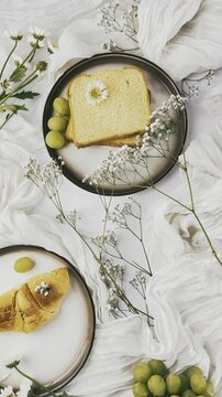 Toasted bread, green grapes, a croissant, pale blossoms, and round plates are arranged among brightly illuminated white fabric with delicate branching stems.