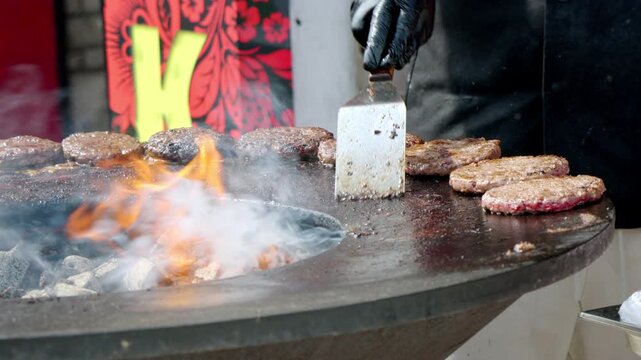 Smoke rises from sizzling burger patties over hot coals. Chef flips each patty with a spatula, expertly. Flames glow beneath a large, round, charcoal grill. The scene suggests outdoor street food