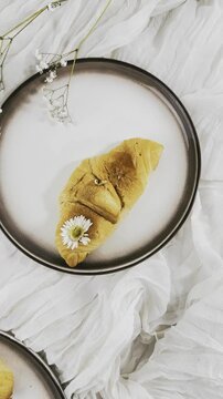 croissant,golden brown pastry,baked goods,flaky pastry,layered crust,browned surface,round plate,dark rim,white flower,yellow center,tiny blossoms,floral stem,branching stem,white fabric,draped fabric