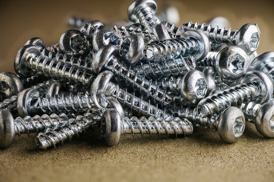 Close-up of a pile of shiny metal screws with Phillips heads arranged on a textured brown surface, showcasing their intricate details and reflections