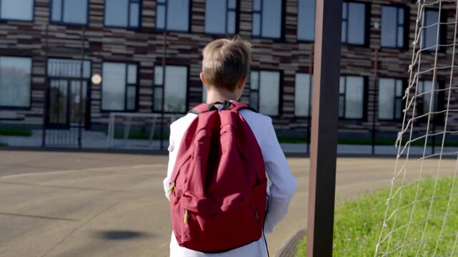 Young boy walks toward school, red backpack prominent. He pauses near a pole, looking at building. Sunlight glints off his hair, soft, natural light. He seems calm, ready for the day ahead