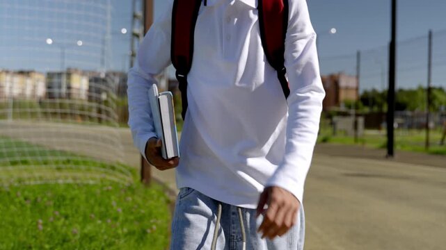 Young boy walks toward school, red backpack prominent. He pauses near a pole, looking at building. Sunlight glints off his hair, soft, natural light. He seems calm, ready for the day ahead