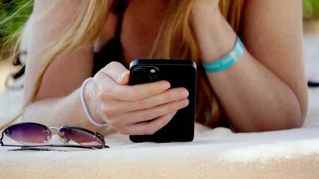 Sunlit beach scene, young woman lying on towel. She holds phone, fingers tapping screen. Glasses rest nearby, worn blue wristband. Relaxed, absorbed in digital world, leisurely mood