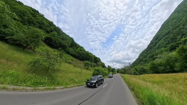 First-person POV driving shot on an asphalt mountain road passing lush green forest hills with a oncoming white car and dramatic cloud formations overhead.