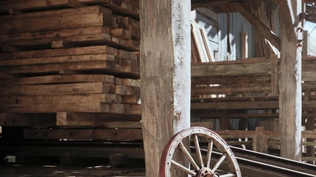 A wooden cart wheel rests against a post close to stacks of lumber in a storage area during daylight hours. The setting shows a rustic and functional space for wood storage.