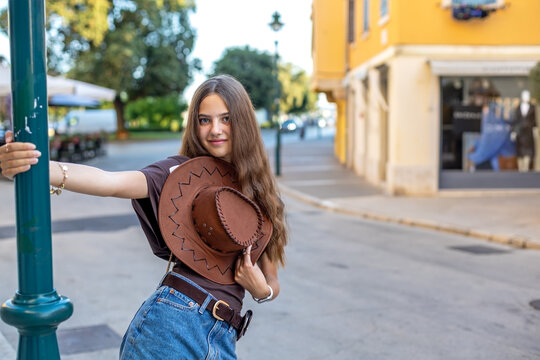 Young Girl Holding Cowboy Hat on City Street. Young girl poses with a cowboy hat while leaning against a street lamp on a quiet city street.