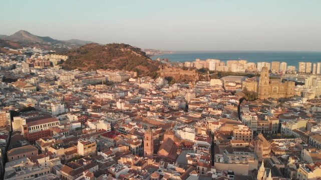 Aerial drone gliding over historic Malaga cityscape at golden hour, revealing ancient cathedrals and terracotta rooftops against the Mediterranean Sea, evoking timeless European charm and cultural dis