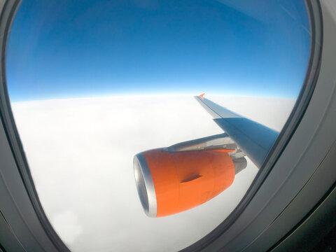 In-flight view from an passenger window looking out at engine cowling and the aircraft wing above the clouds. Curved plastic frame of the window the bright sky outside. Blue sky, white cloud layer. 