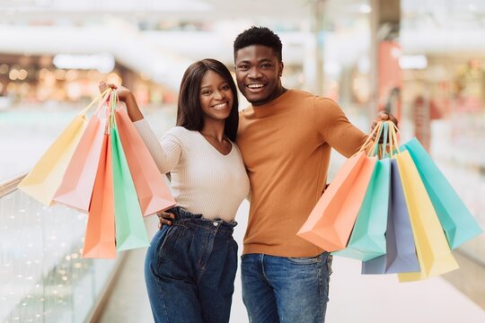Happy Black Couple Smiling While Shopping Together, Holding Colorful Shopping Bags In A Modern Mall, Enjoying Their Shopping Experience. Cheerful Young Adults Having Fun At Retail Store.