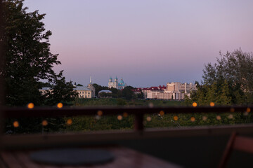 Naklejka premium Scenic twilight view over Grodno, Belarus city skyline, featuring St. Francis Xavier Cathedral and Regional Drama Theatre, viewed through a railing with warm bokeh lights at dusk from a terrace.