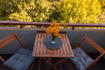 Naklejka premium Cozy balcony setting featuring a wooden table with bright yellow flowers in a vase, two folding chairs with cushions, and a scenic view of autumn trees in warm sunlight.