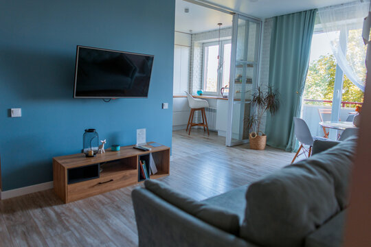 Stylish modern living room interior with blue wall, mounted TV, wooden console, comfortable sofa, and bright open plan layout connected to kitchen and balcony.