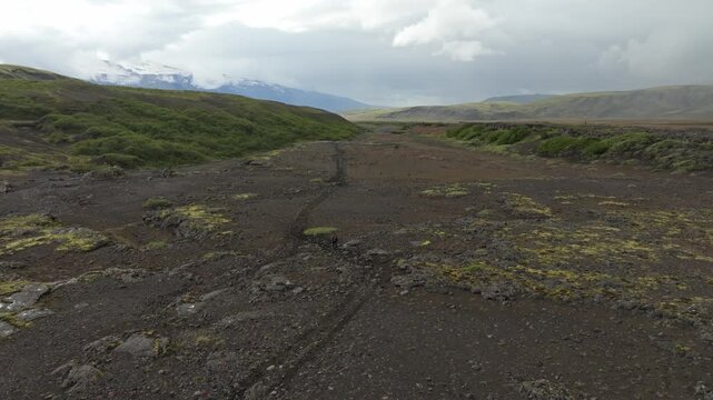 Drone pushes in on a lone hiker traversing the rugged Laugavegur trail, showcasing Iceland's dramatic volcanic landscape under an overcast sky.