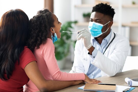 Focused Young Black Girl With Mother Present Having Throat Exam By Doctor In Pediatric Office. Child Patient Engaged During Consultation With Healthcare Professional In Friendly Setting.