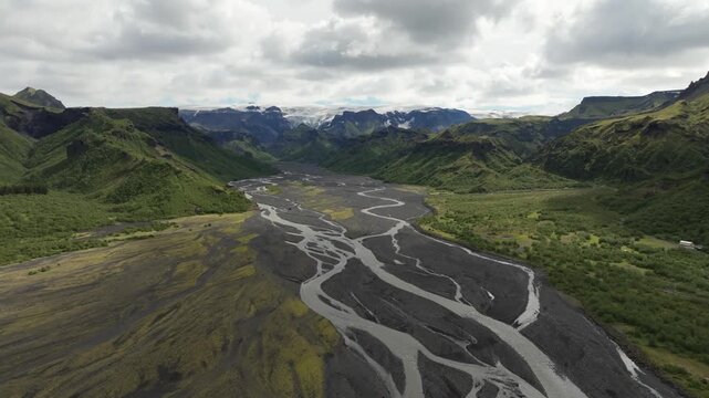 A drone shot pushes in and booms up over the glacial valley in &THORN;&oacute;rsm&ouml;rk, Iceland, showcasing a vast glacial river valley. Majestic mountains and a distant glacier frame the rugged, green landscape.
