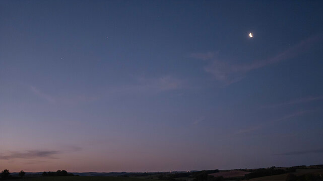 Twilight sky with a delicate crescent moon and faint wisps of clouds, transitioning from deep blue to soft purple, over a dark, silhouetted rolling landscape.keywords: twilight, dusk, evening, sky