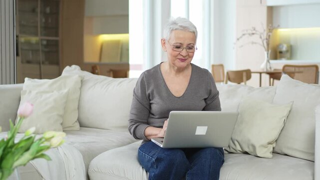Confident stylish happy middle aged senior woman using laptop at home. Stylish older mature 60s grandmother sitting at couch looking at computer screen typing chatting reading writing email