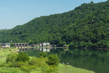 Fototapeta premium Tranquil reservoir at Jinni Dam in Toyama, Japan, framed by lush green mountains with calm reflective water on a clear summer day.