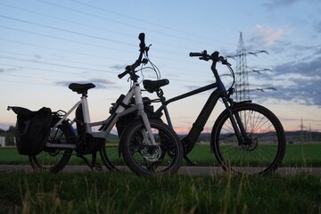 Two electric bicycles stand beside a grassy field in Germany, with power transmission lines, a lattice tower and electrical infrastructure visible at dusk, expressing a theme of electrification © Oli-Eugen