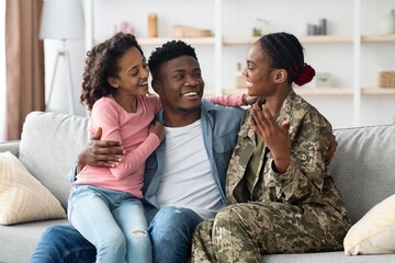 Obraz premium Smiling African American Woman In Military Uniform Embracing Husband And Daughter At Home, Enjoying Family Time Together On Couch, Exuding Happiness And Love, Family Concept.