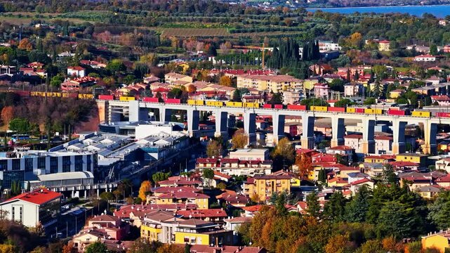Aerial view of a freight train crossing a concrete viaduct over a residential town with autumn trees near Lake Garda, Italy.