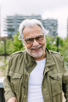 Happy senior man wearing stylish eyeglasses and an olive green jacket, confidently smiling at the viewer while enjoying leisure time outdoors in a bustling urban environment