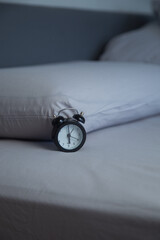 On the light gray bed linen there is a black miniature alarm clock against the background of two soft pillows in the dim lighting. Concept: the feeling of early morning, calm bedroom interior. High © Ксения Мнасина