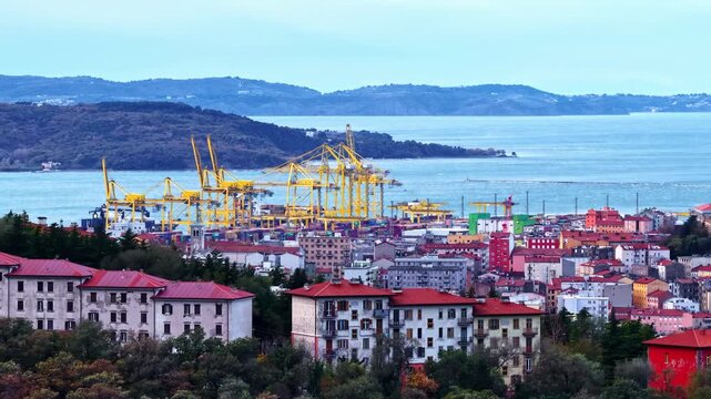 Aerial view of the busy Port of Trieste with yellow industrial cranes, shipping containers, and urban buildings along the Adriatic coastline in Trieste, Friuli-Venezia Giulia, Italy.
