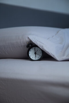 Vertical photo of a black alarm clock lying on the bed next to a pillow, partially covered with the edge of a gray blanket on a gray background. The concept of getting up in the morning, sleeping