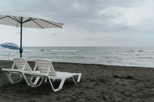 Empty white sun loungers and umbrella on a dark sand beach under a cloudy sky