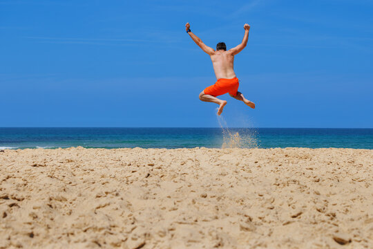 Excited man on vacations jumping above sandy beach beside ocean