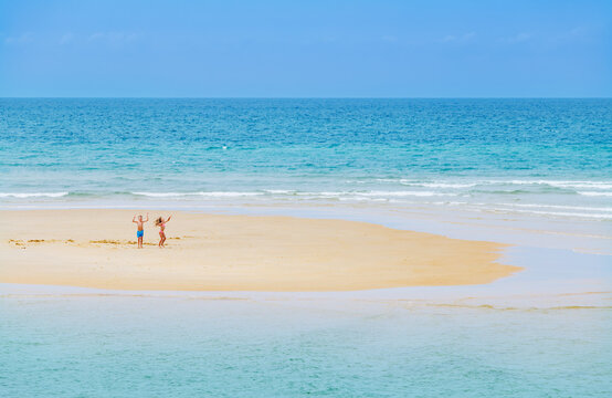 Two kids wave and jump on a sandy island alone in blue ocean