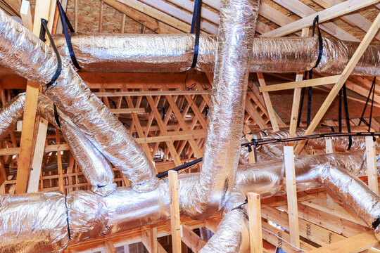 Workers are installing metal ductwork in attic of construction site, wooden beams support structure.